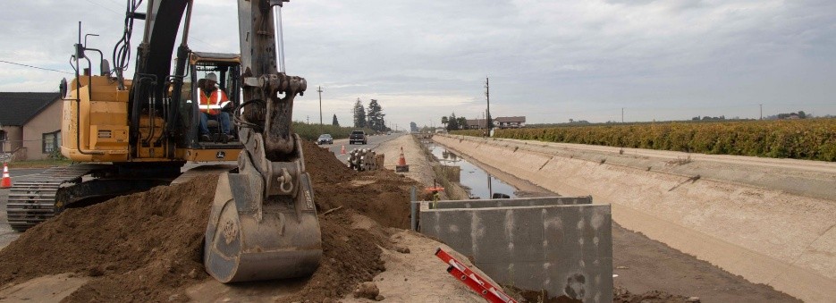 A construction site featuring an excavator digging near a road, with a drainage ditch and farmland in the background.